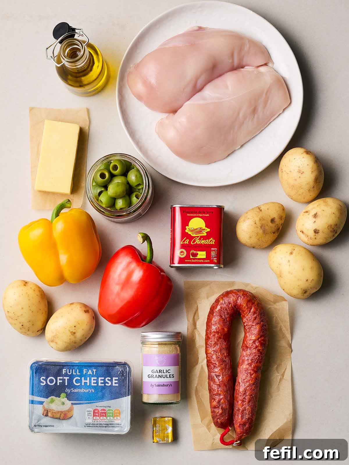 Raw ingredients neatly arranged on a pristine white counter, featuring fresh chicken breasts, whole potatoes, a ring of chorizo, vibrant bell peppers, a tub of soft cream cheese, and various spices, ready for preparation.