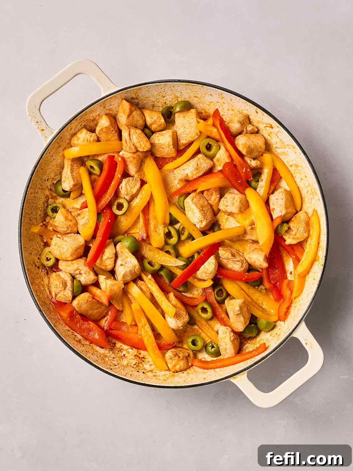A pan filled with diced chicken, colorful sliced peppers, and green olives, actively cooking in the residual chorizo oil, creating a fragrant and inviting mixture.