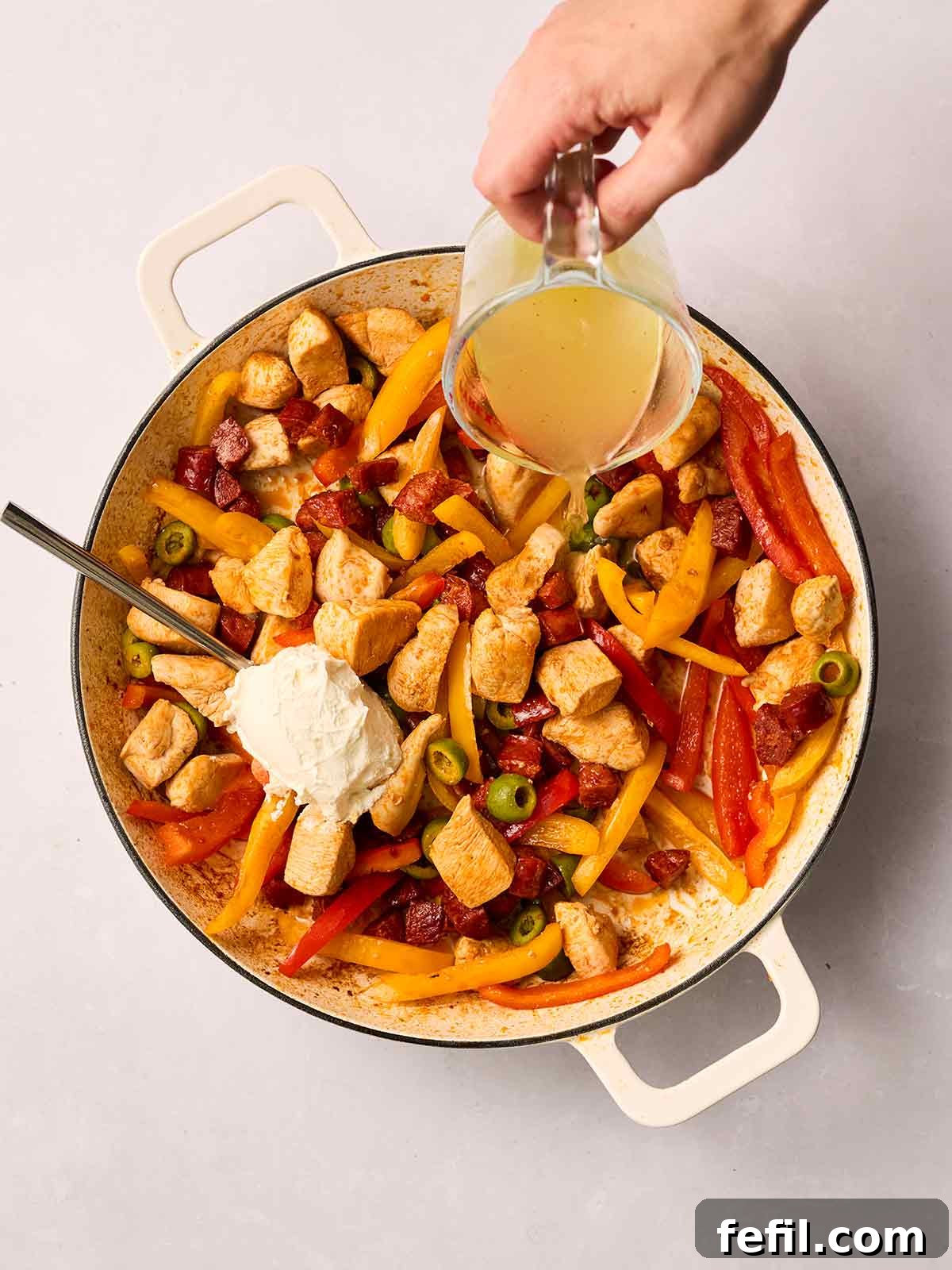A skilled hand pours hot chicken stock into the pan of chicken, chorizo, and vegetables, while a spoon stirs in creamy white cream cheese, creating a rich and inviting sauce.