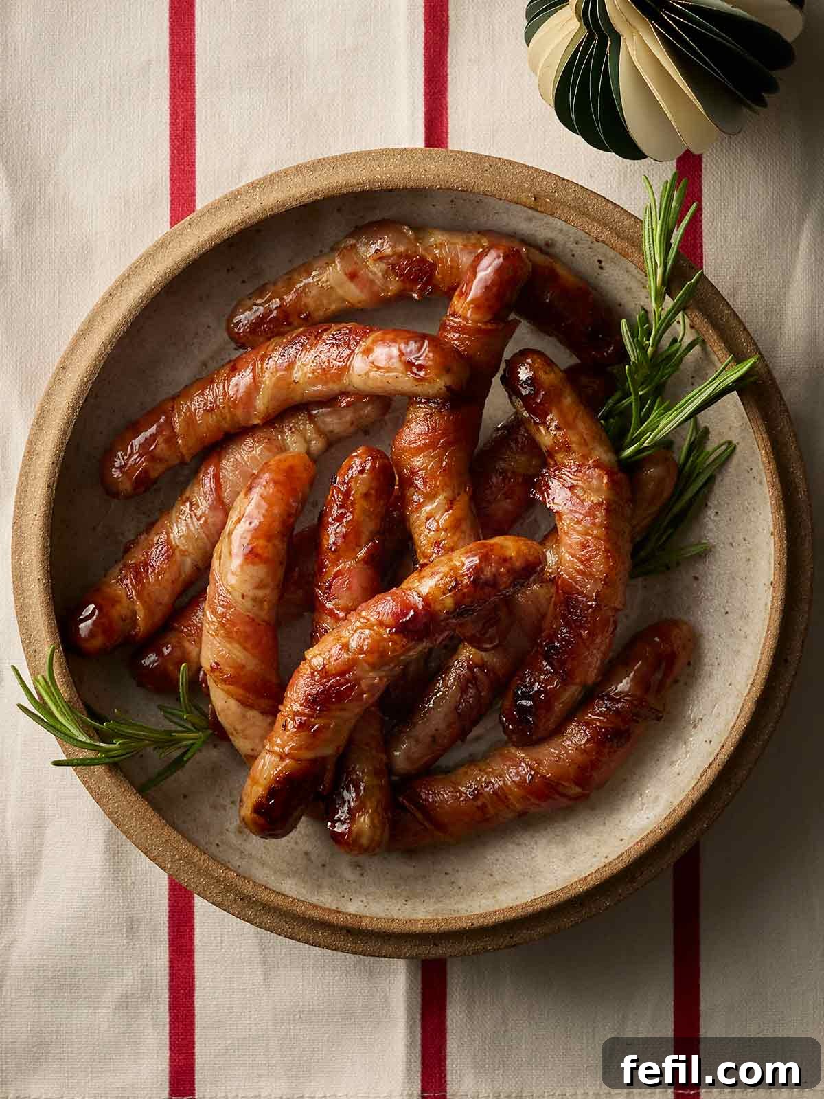 A plate of perfectly cooked Pigs in Blankets, garnished with fresh rosemary, resting on a charming red and white striped tablecloth, ready for festive feasting.