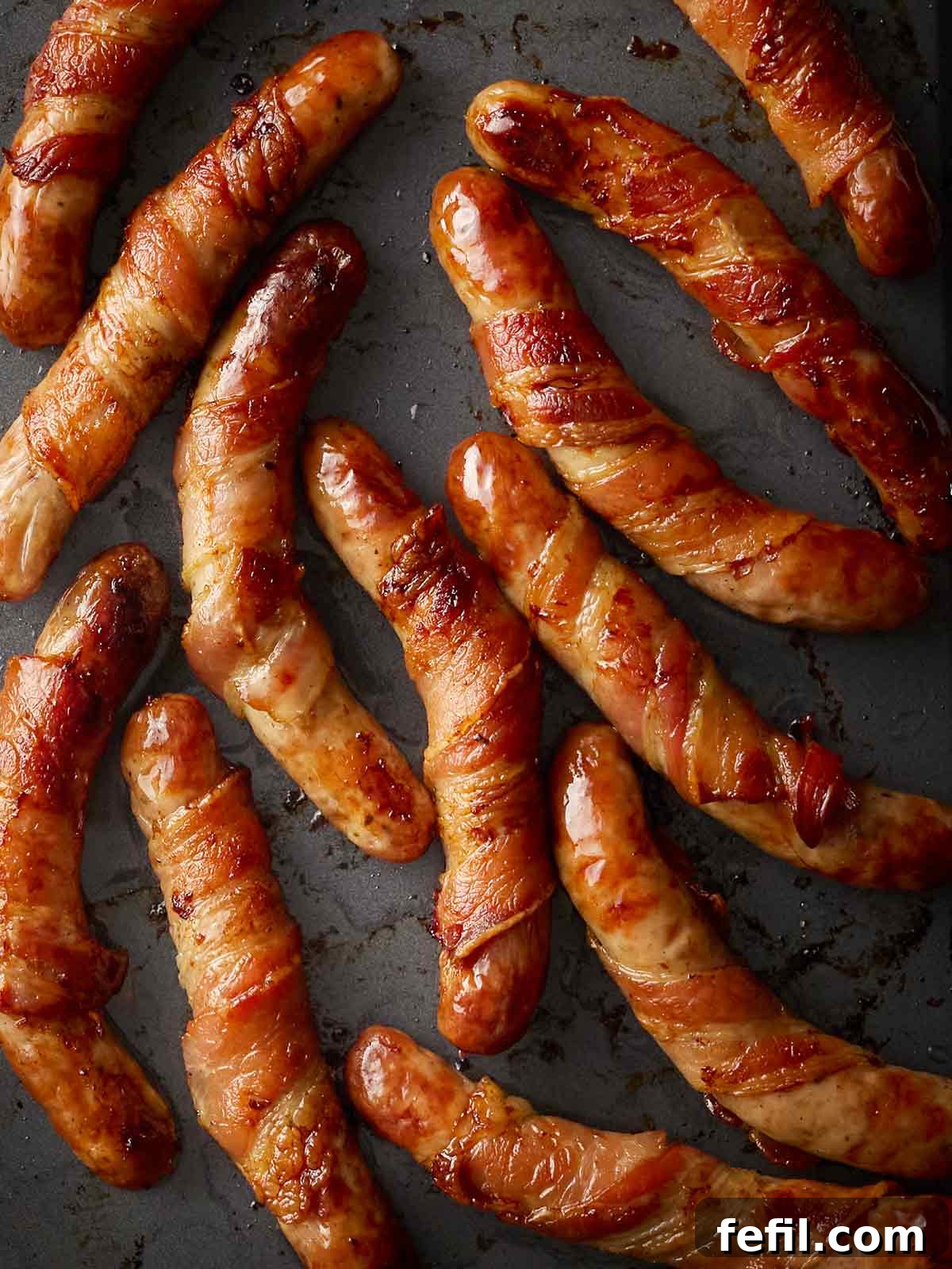 Raw Pigs in Blankets arranged on a black oven tray, ready for baking to golden perfection.