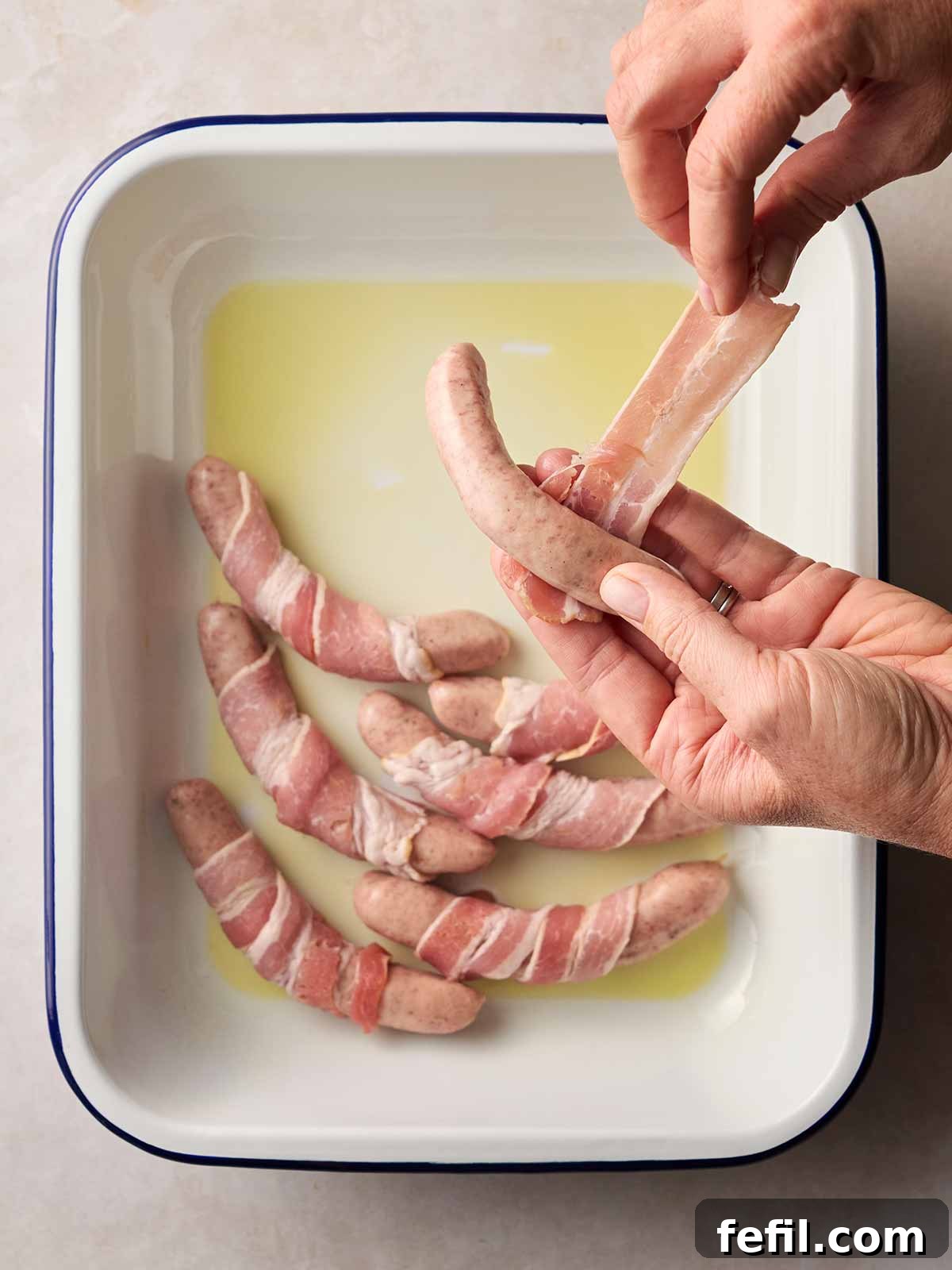 A baking dish with a thin layer of oil, surrounded by prepared bacon-wrapped sausages, with a hand actively wrapping a raw sausage in a strip of bacon.