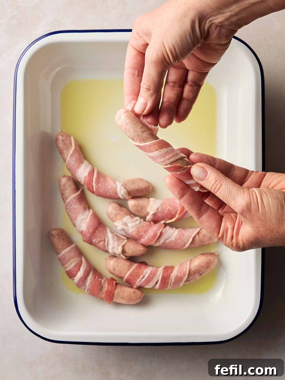 Two hands skillfully wrapping strips of bacon around chipolata sausages, with a prepared baking dish in the background, ready for more Pigs in Blankets.