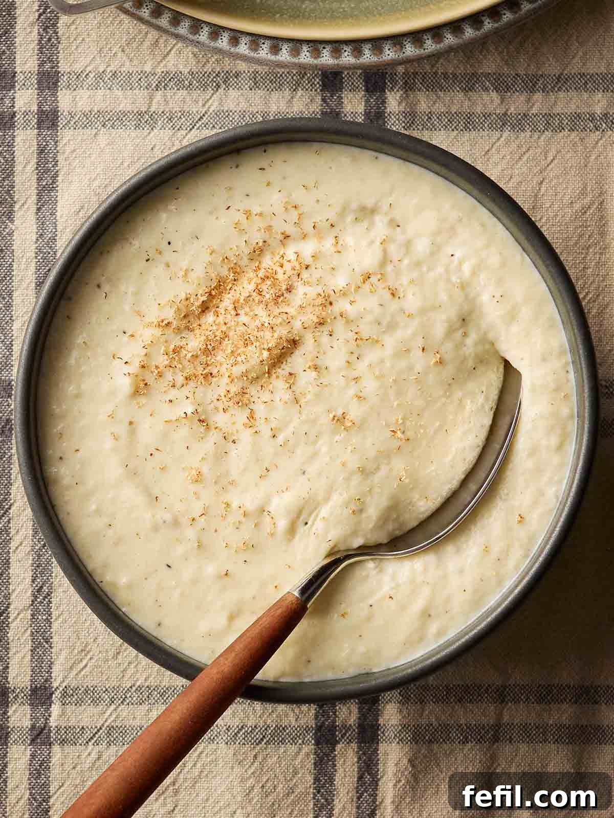 A beautiful bowl of creamy homemade Bread Sauce, ready to be served, resting on a festive checked tablecloth with a spoon dipped inside.