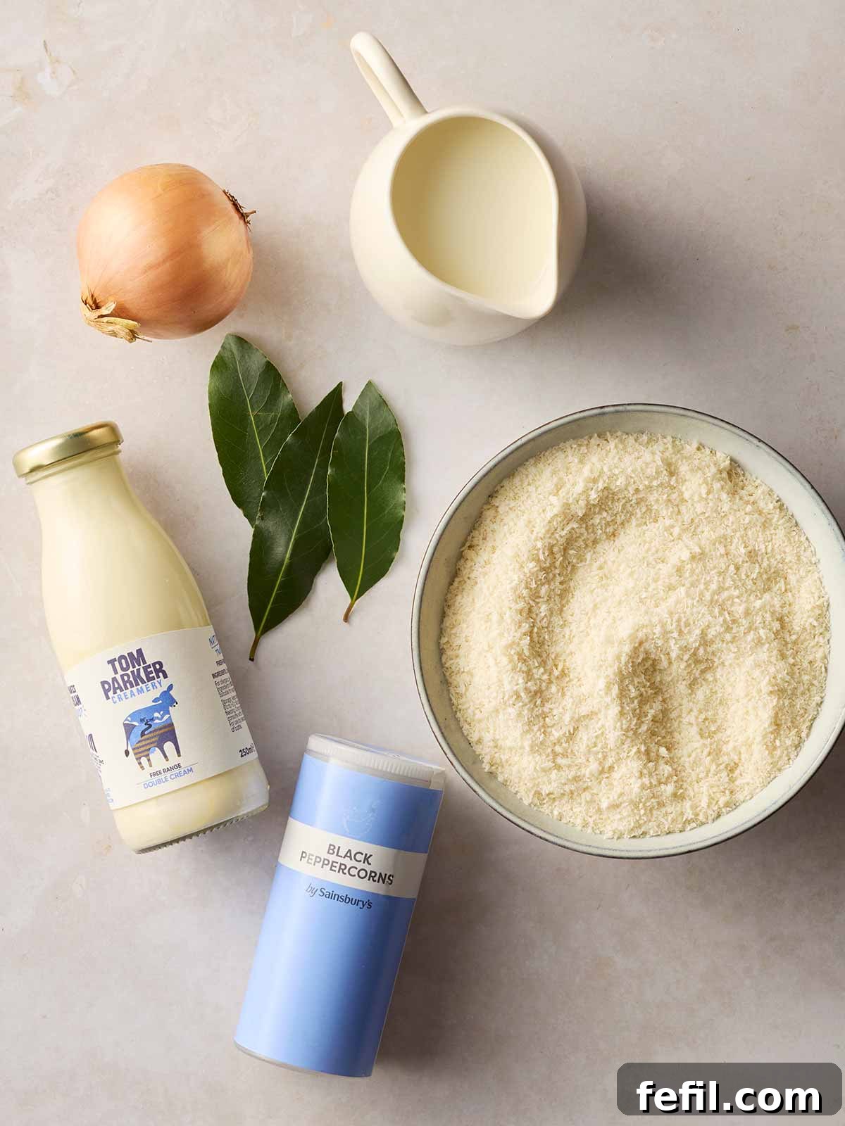 A pristine white counter showcases the essential ingredients for homemade Bread Sauce: fresh breadcrumbs, creamy milk, aromatic bay leaves, a quartered onion, whole black peppercorns, and rich double cream.