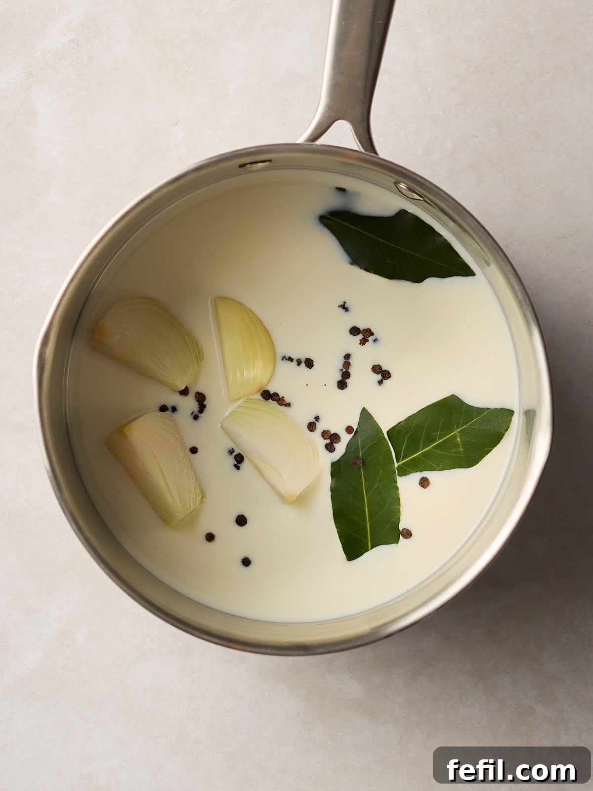 A saucepan on the stove filled with milk, quartered onion, bay leaves, and whole peppercorns, gently infusing.