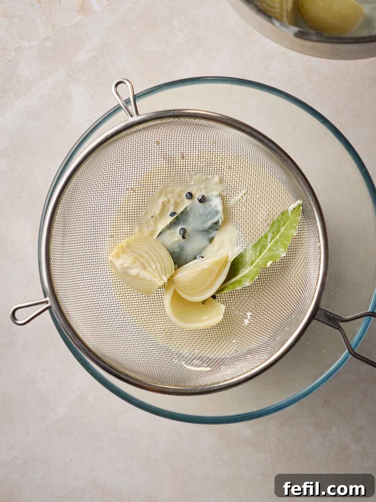 A sieve positioned over a glass bowl, capturing the infused onion and bay leaves, while the spiced milk drains into the bowl below.