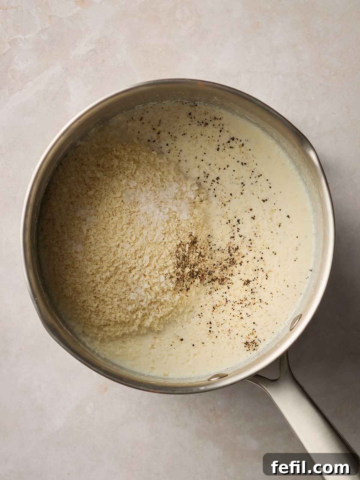 A saucepan containing the strained, spiced milk, now with breadcrumbs sprinkled on top, ready to be stirred and thickened into Bread Sauce.