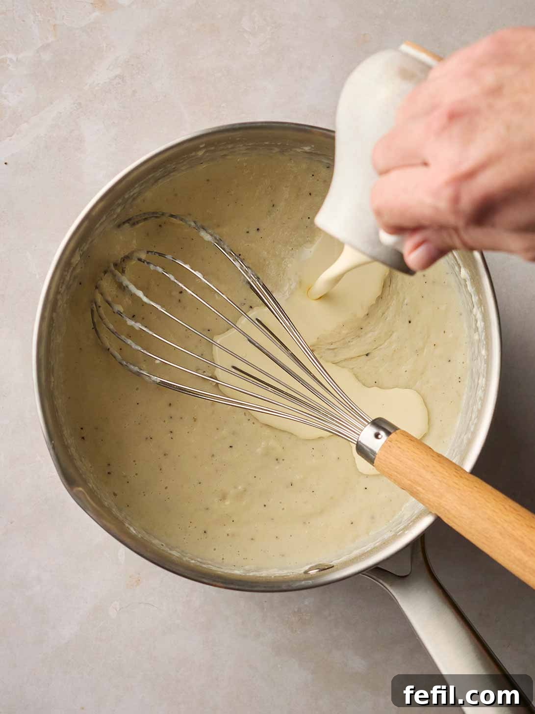 A hand pouring a stream of rich double cream from a jug into a pan of thickening Bread Sauce, with a whisk nearby.