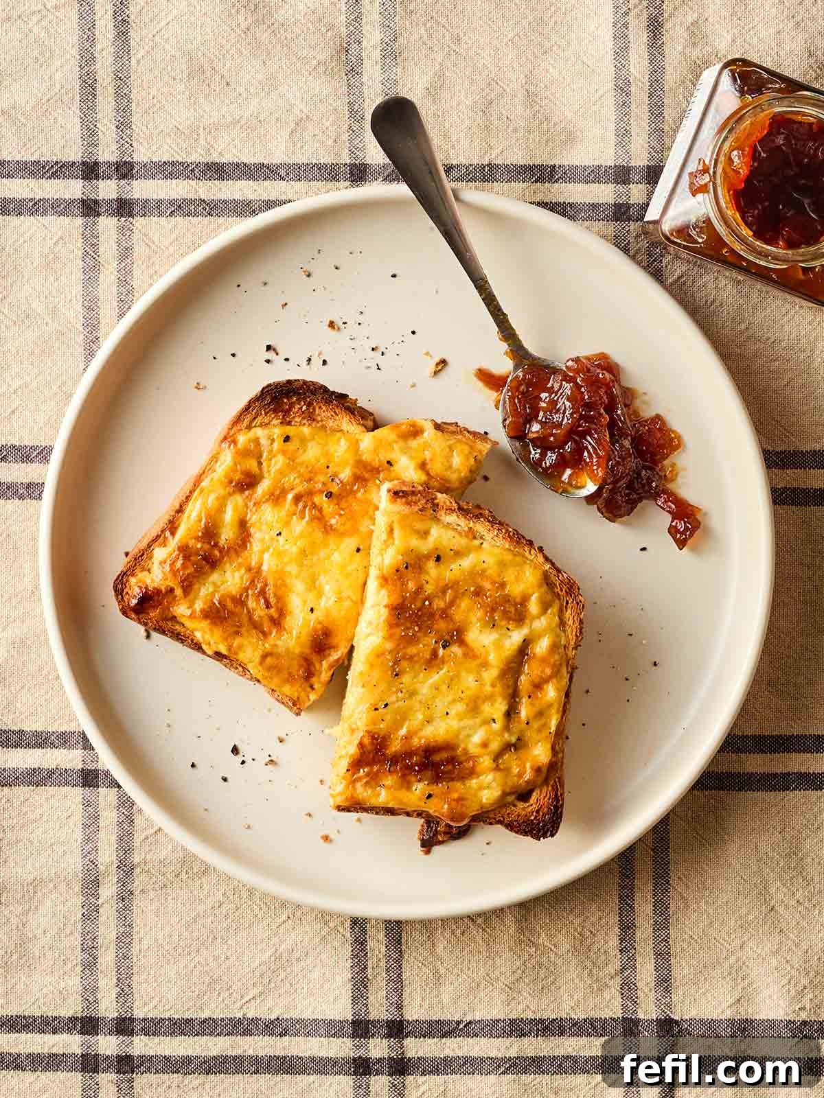 A plate with a slice of Welsh rarebit with a spoonful of chutney to the side, on a checked tablecloth.