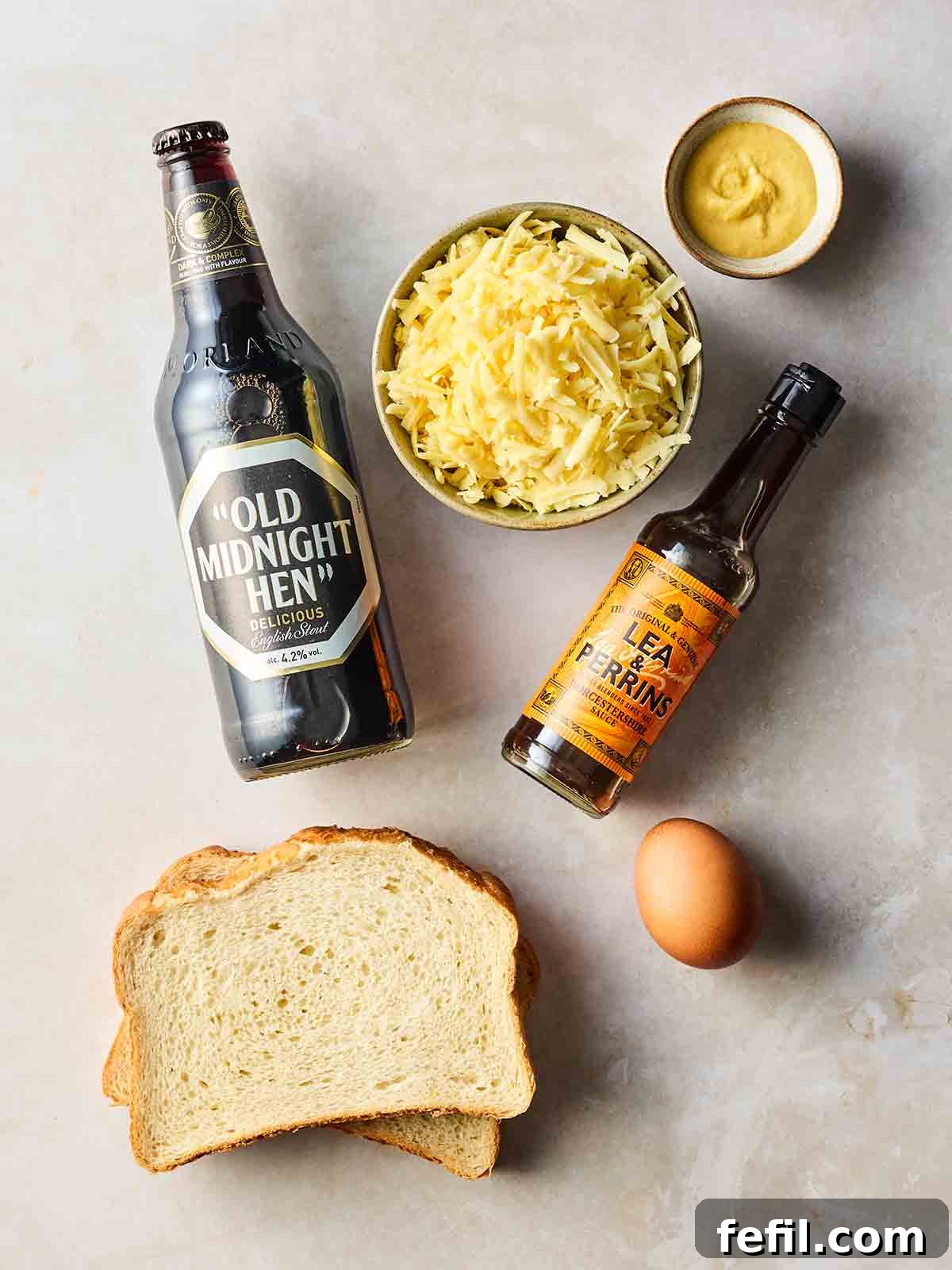 The ingredients for making Welsh rarebit laid out on a white surface, including cheese, an egg, beer, mustard, and Worcestershire sauce.