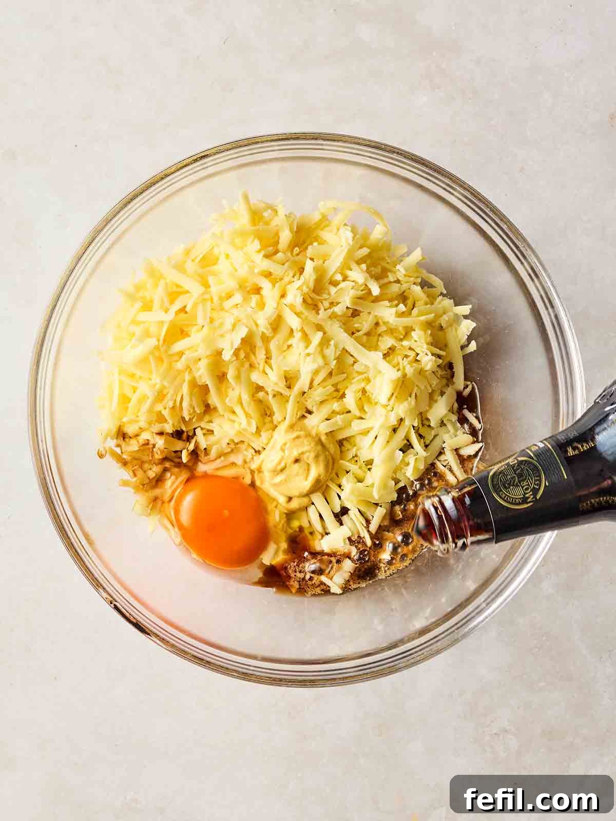 A glass bowl filled with grated cheese, an eggs and some beer being poured in, demonstrating the mixing of rarebit ingredients.