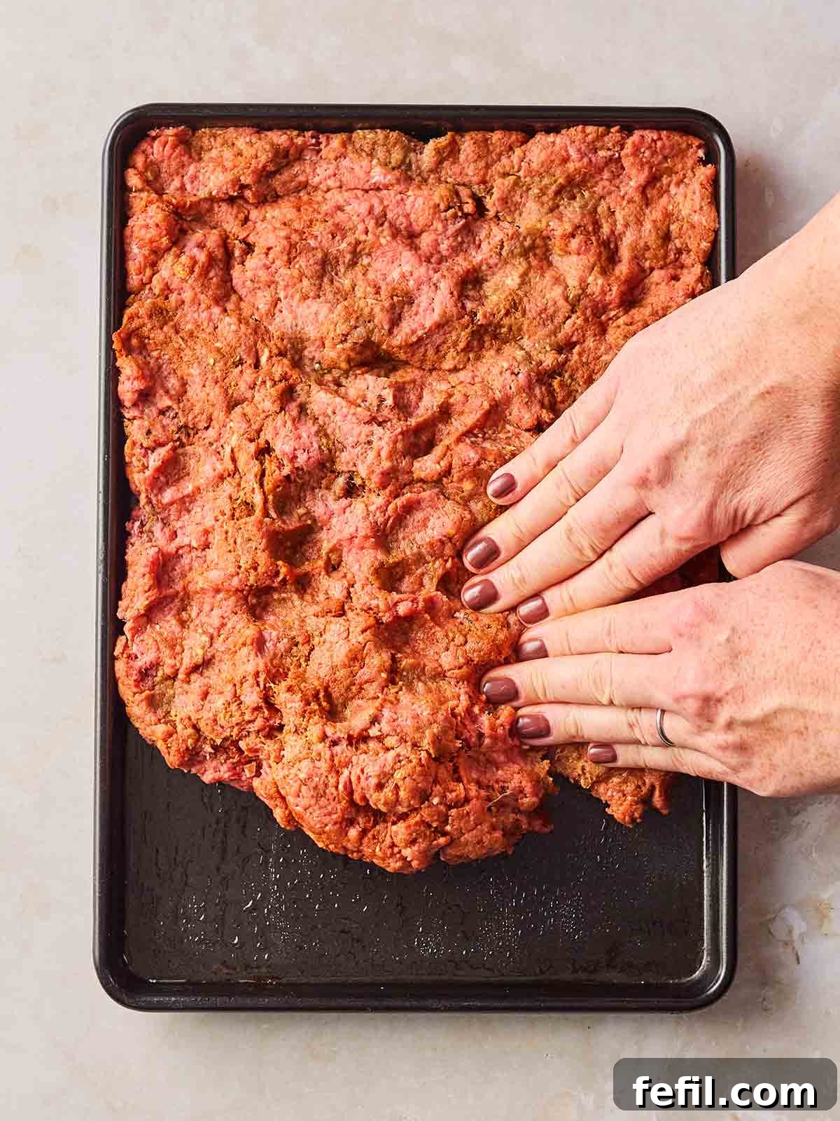 Two hands pressing a beef mince mixture into an oven dish for a meat sheet kebab recipe.