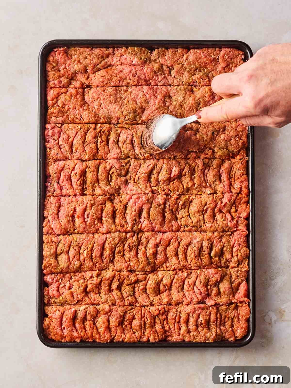 An oven sheet with a mince mixture pressed into it, and a hand scoring the meat with a spoon.