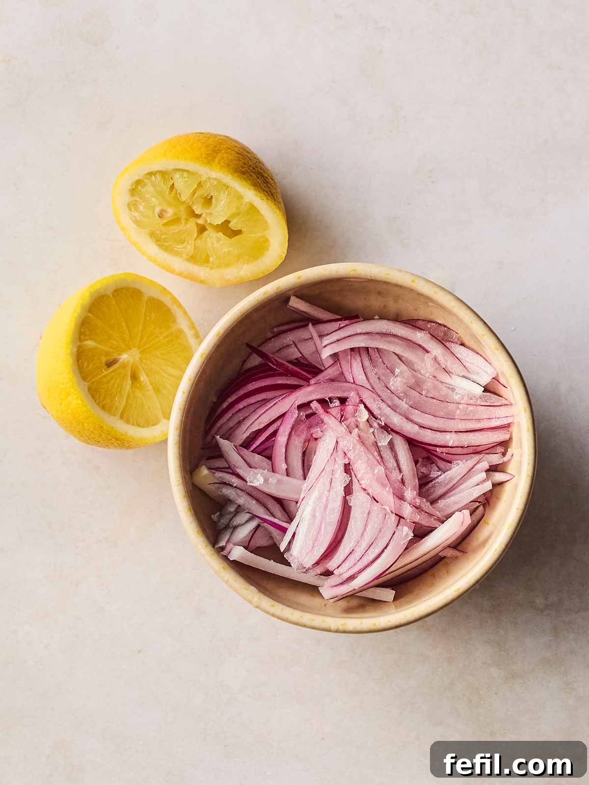A sliced red onion in a dish with salt over and an empty half a lemon on its side next to it, showing it has been squeezed into the onion dish.