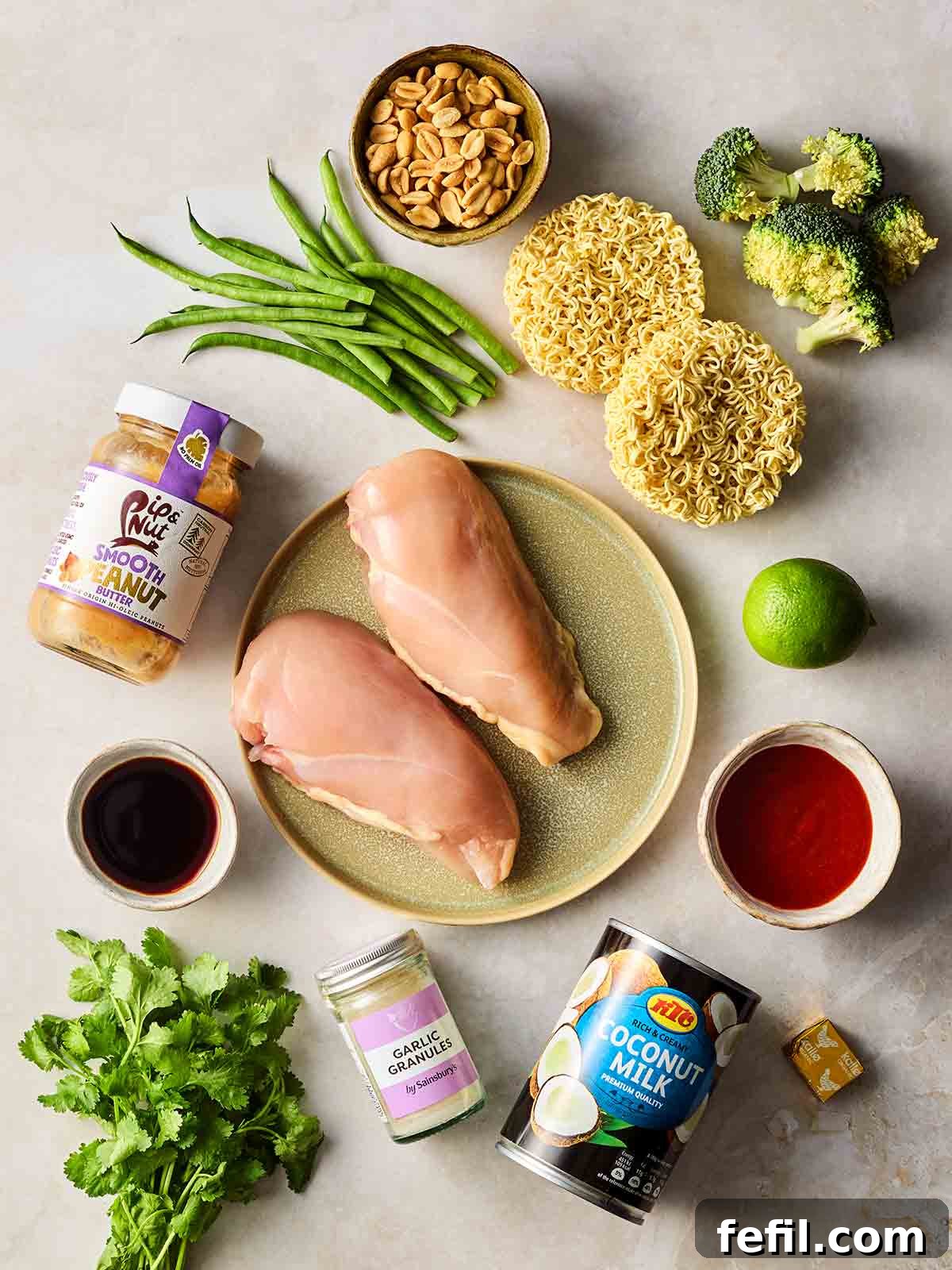 Spicy Peanut Chicken Noodles 4 Ingredients laid out on a white surface, including raw chicken breasts, noodles, vegetables, coconut milk, peanut butter and more.