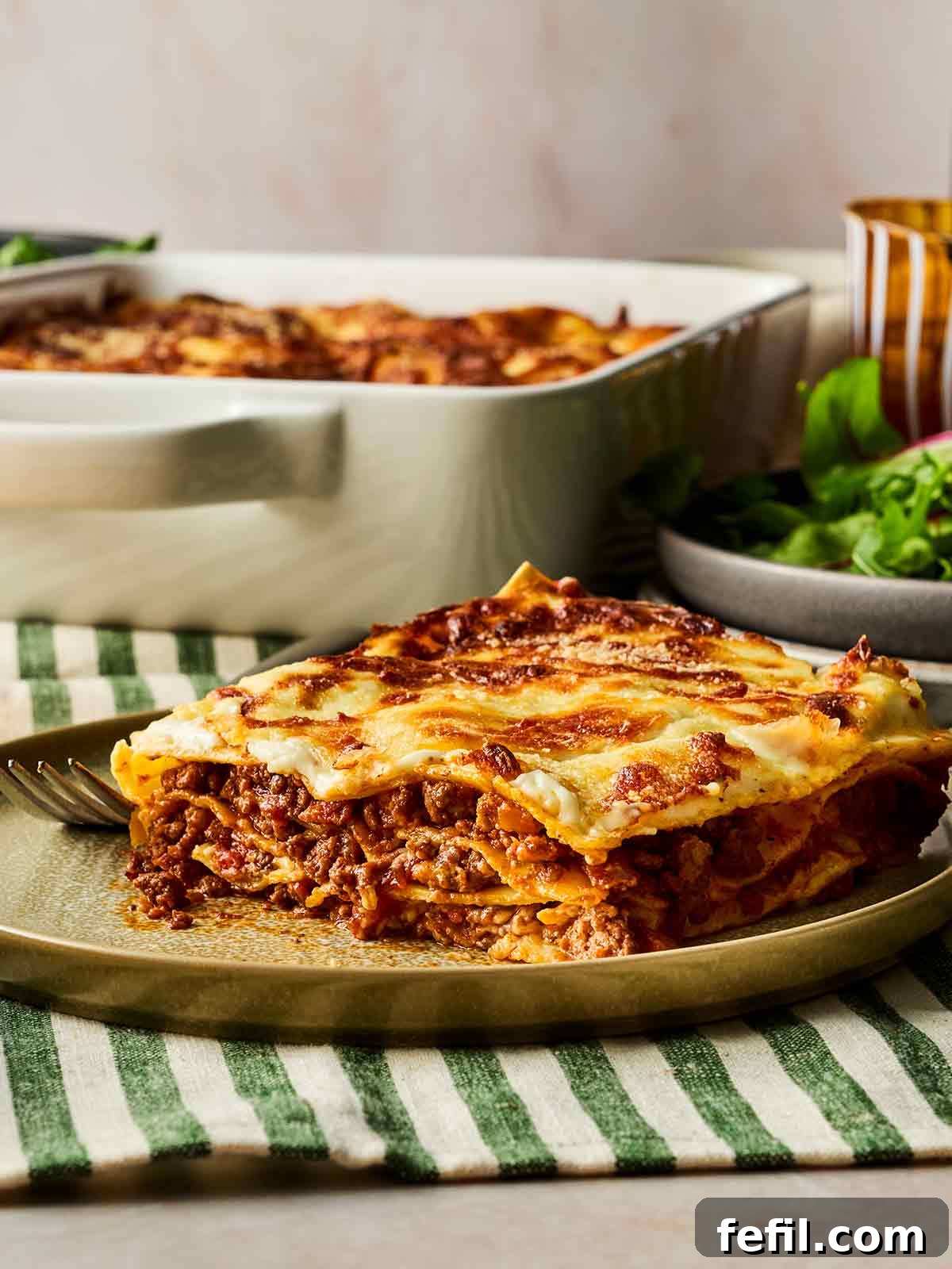 The Ultimate Classic Lasagne 2 A plate in the foreground with a portion of lasagne on, in the background the oven dish with the rest of the lasagne, on a dining table.