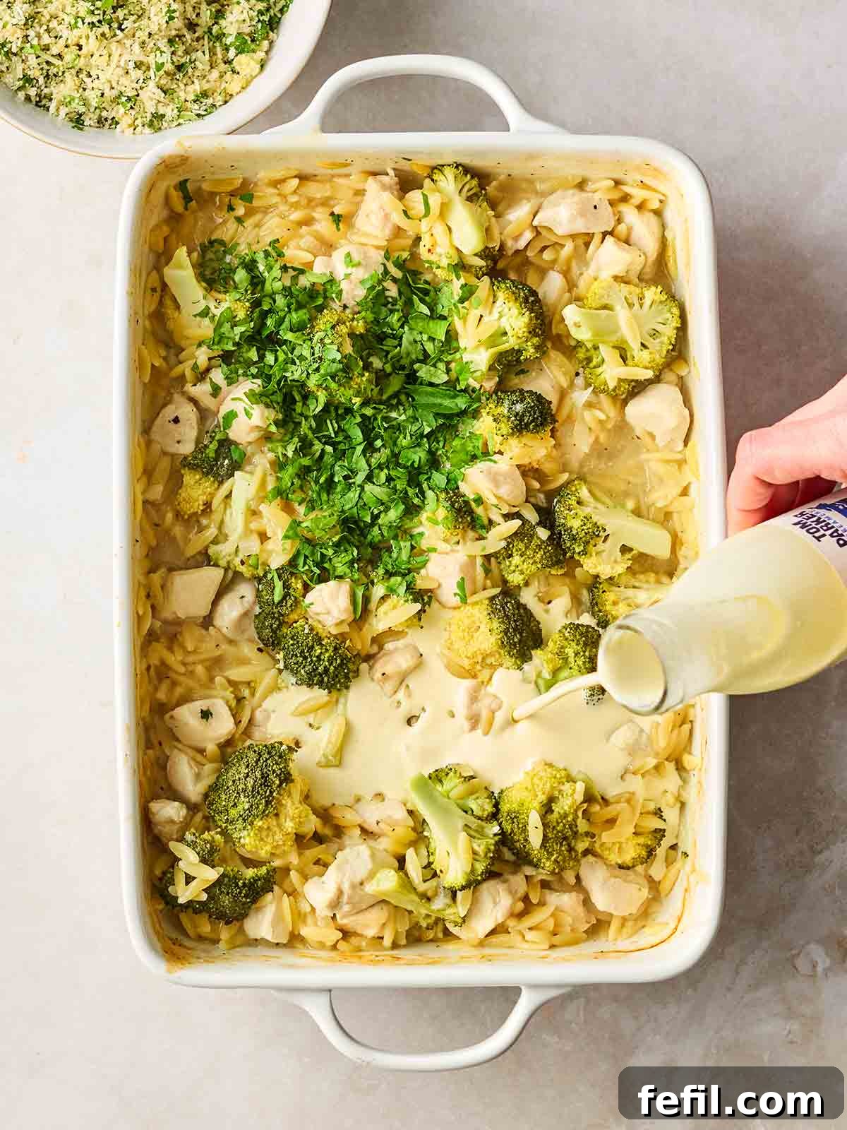 Herb Butter Chicken Pasta 8 An oven dish filled with cooked chicken, orzo pasta, and broccoli, with a hand pouring in rich double cream, ready for the final steps before grilling.