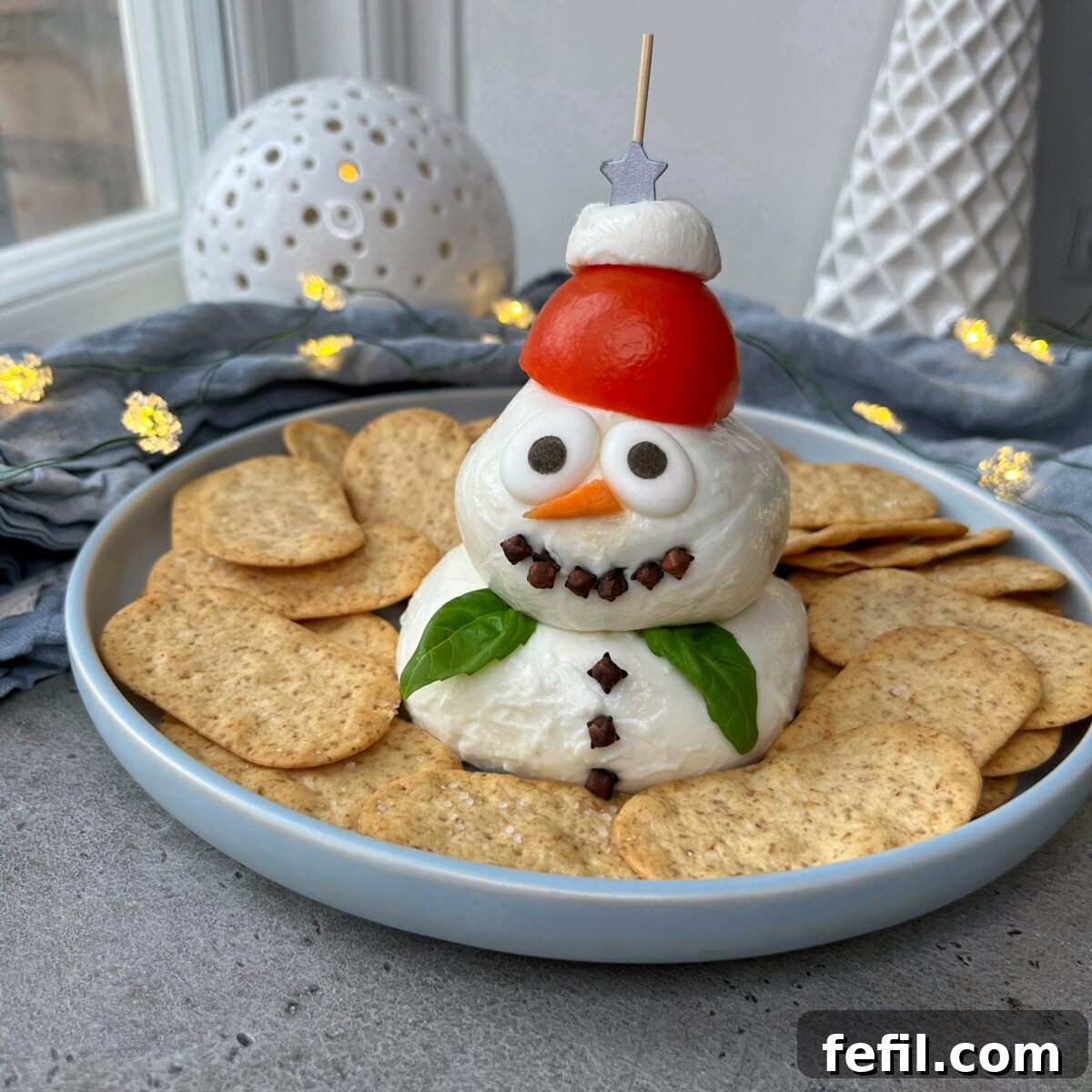 A Very Merry Cheese Board 2 A snowman-shaped cheese ball decorated with a tomato hat and cloves, surrounded by crackers on a plate.