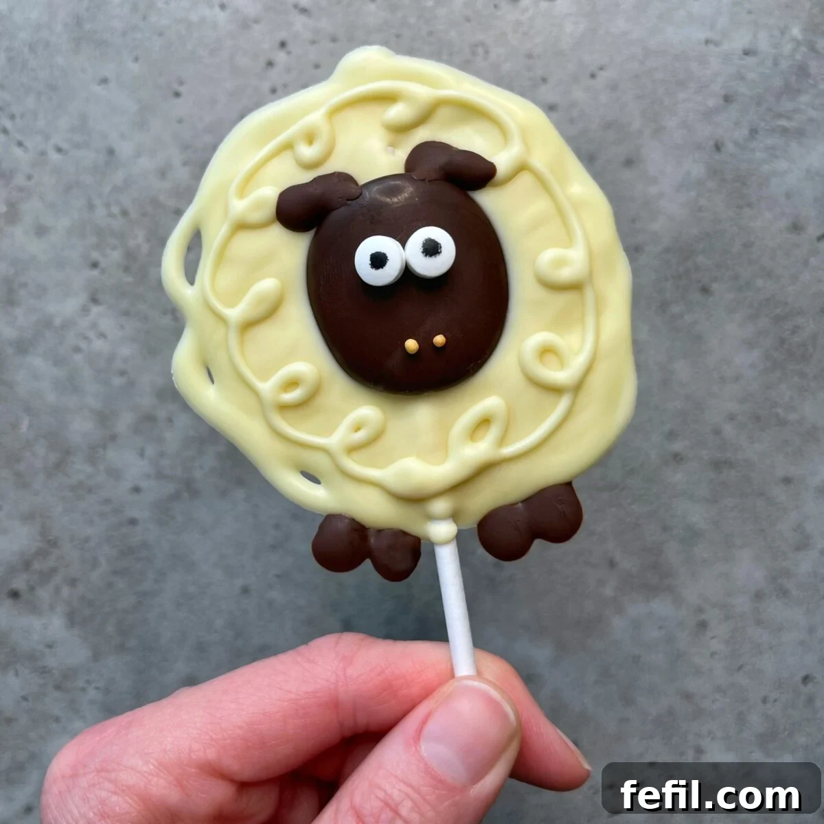 A hand delicately holds a single, enchanting sheep-shaped lollipop. It features a rich dark chocolate face, contrasted by bright white chocolate eyes, and a beautifully swirled white chocolate body, giving it a fluffy appearance. This adorable bite is perfect for creative food recipes, showcased against a soft gray background.