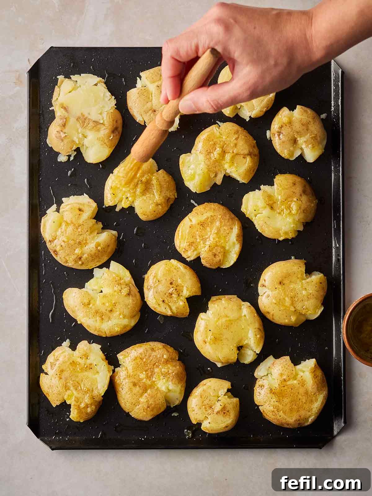 A baking tray with smashed potatoes being brushed with olive oil by a hand.