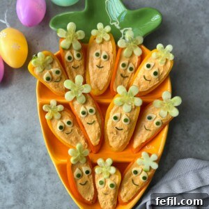 A carrot-shaped tray holds health-inspired orange carrot boats with dip, decorated with cucumber eyes, small green smiley faces, and celery flower tops. Colorful Easter eggs are nearby on a gray surface.
