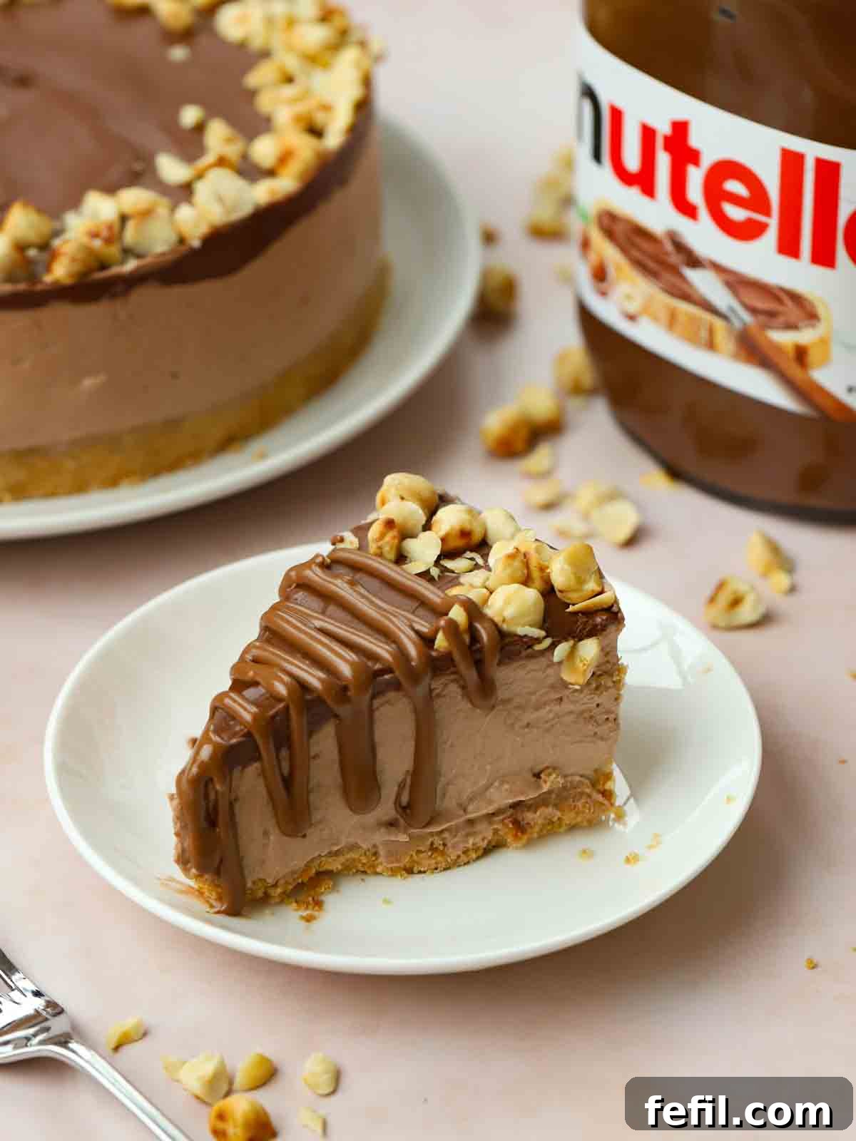 A chocolatey slice of No-Bake Nutella Cheesecake on a plate, ready to eat, with the rest of the bake in the background, showing off its layers.