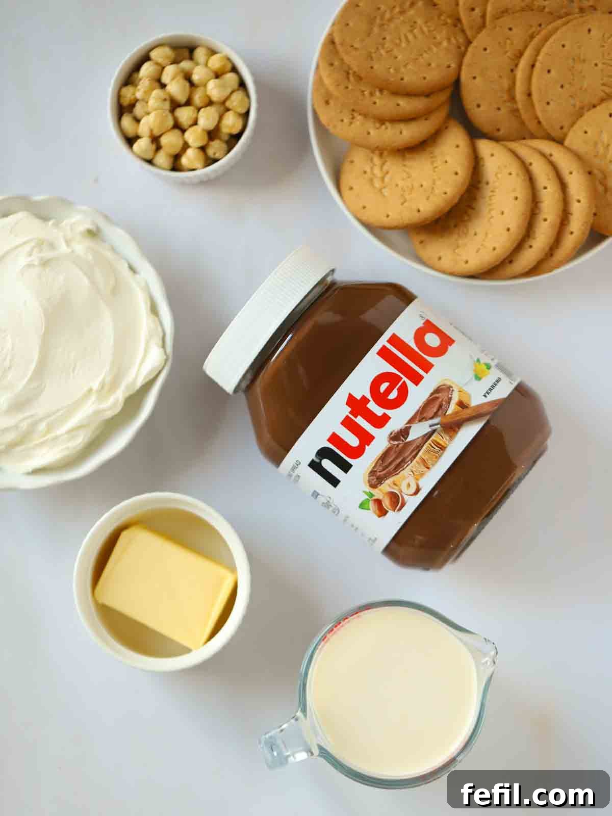 The ingredients to make a Nutella Cheesecake laid out on a white counter, including biscuits, butter, cream, cream cheese, Nutella, and hazelnuts.