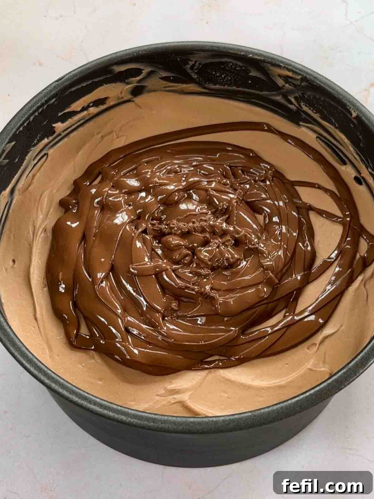 A baking tin filled with whipped cream mixture and topped with chocolate ready to spread for for step 5 in the recipe for Nutella Cheesecake.
