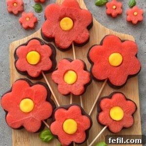 Sliced watermelon and yellow fruit arranged as flower shapes on sticks, resembling lollipops, displayed on a wooden board with small fruit flowers, basil leaves, and chocolate fruit skewers for a delightful touch.