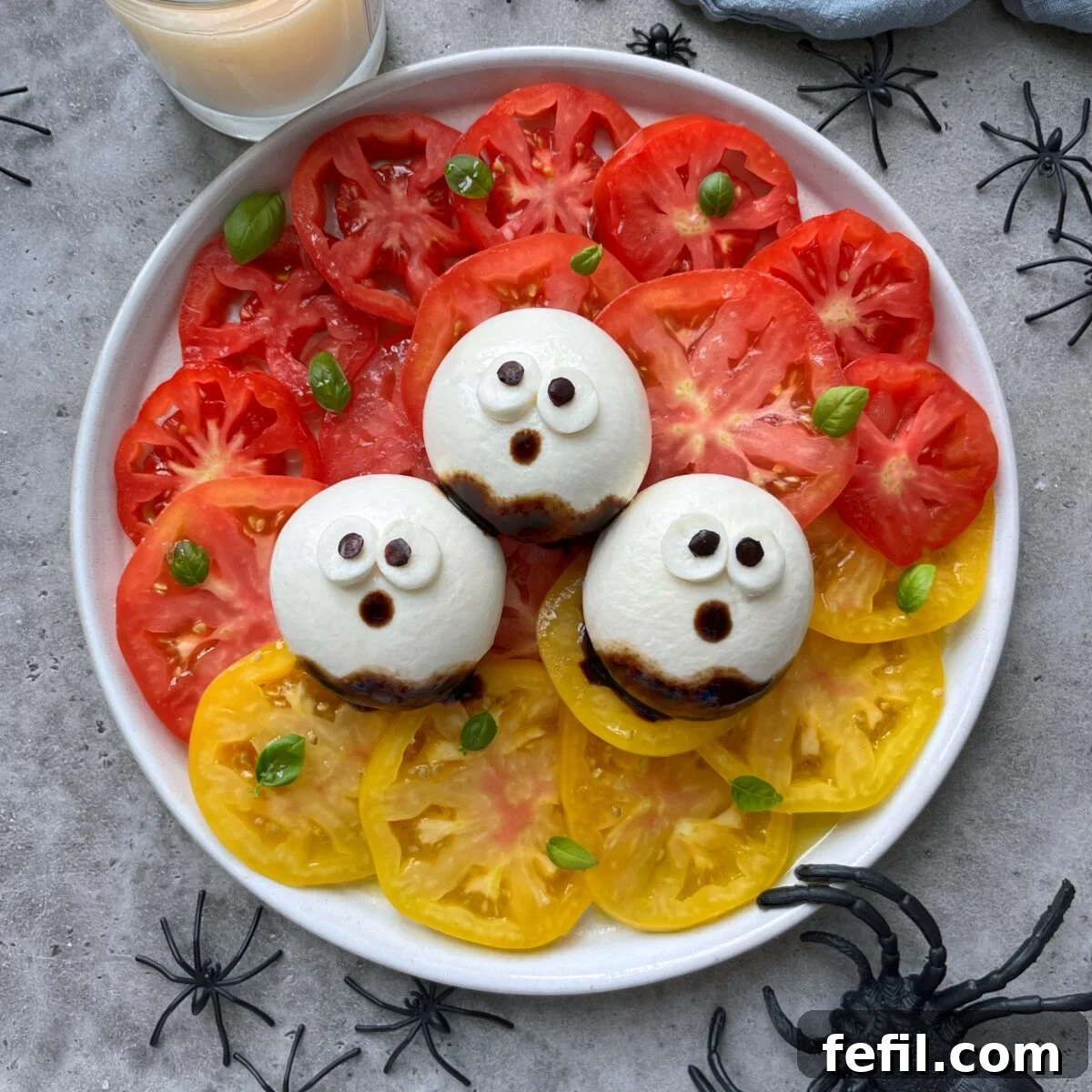 A plate of sliced red and yellow tomatoes topped with three burrata balls decorated as ghost faces, surrounded by plastic spiders.