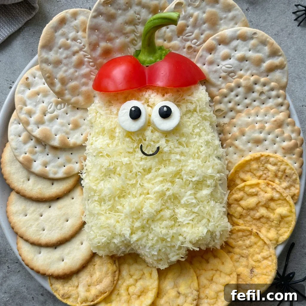 A Halloween-themed cheese ball shaped like a ghost, with shredded cheese, olive eyes, red bell pepper for a hat, and a smiling face. It's served with assorted crackers on a plate.