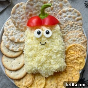 A Halloween-themed cheese ball shaped like a ghost, with shredded cheddar cheese, olive eyes, red bell pepper hat, and a smiling face. Surrounding it are assorted crackers.