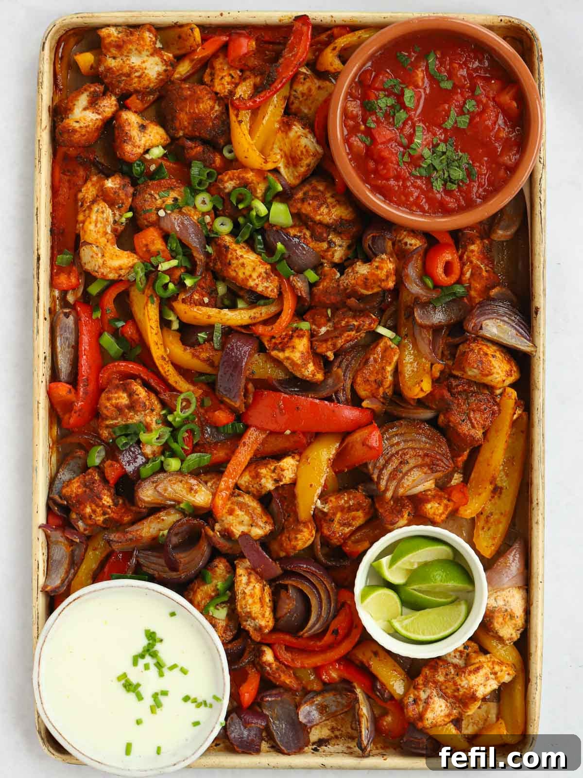 A tray with oven baked chicken fajitas, with side dishes in bowls of salsa, sour cream and lime.
