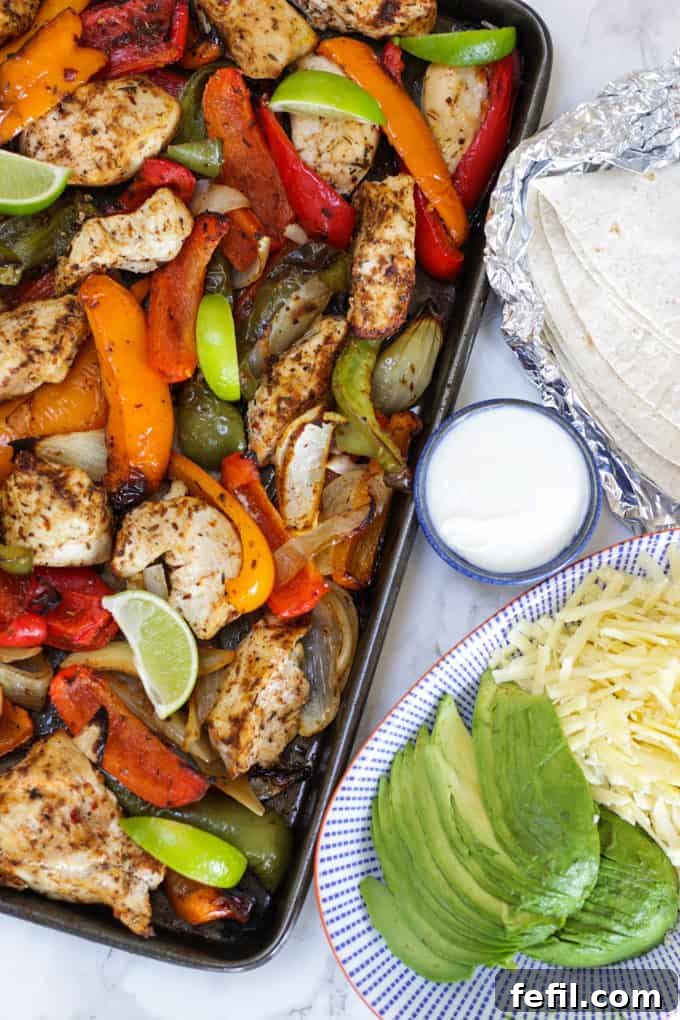 Oven baked chicken fajitas on a baking tray with peppers and lime wedges and avocado and cheese on a blue striped plate.