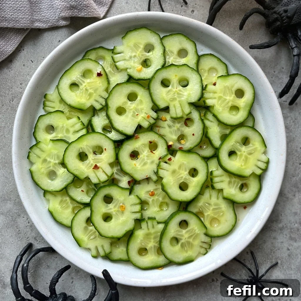 Chilling Cranium Cucumbers 2 Cucumber slices carved to look like skulls are arranged on a white plate, sprinkled with red pepper flakes for a healthy twist. Plastic black spiders surround these festive bites, making them perfect for Halloween-themed food recipes.