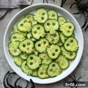 Cucumber slices carved to look like skulls are arranged on a white plate, sprinkled with red pepper flakes for a healthy twist. Plastic black spiders surround these festive bites, making them perfect for Halloween-themed food recipes.