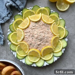 A round plate of creamy Hot Smoked Salmon Dip is garnished with alternating slices of lemon and cucumber in a circular pattern. A blue napkin, lemon halves, and toasted bread slices are nearby on a gray surface.