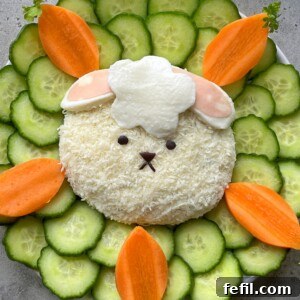 A cute Easter Cheese Ball shaped like a lamb, topped with white cheese and sliced mortadella, is surrounded by cucumber slices and carrot pieces arranged in a flower pattern on a white plate.