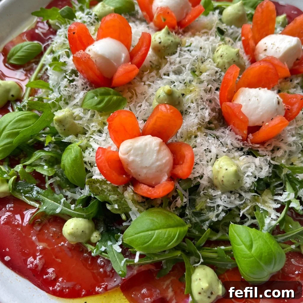 A beautifully plated Beef Carpaccio Salad with thin beef slices, crisp arugula, generous shavings of Parmesan, creamy avocado dressing dollops, and artfully carved tomato-bocconcini flowers, all garnished with fresh basil.