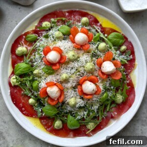 A vibrant Beef Carpaccio Salad features thin tomato slices, arugula, basil leaves, avocado cream dollops, grated cheese, and mozzarella balls with carrot flowers. All are drizzled with olive oil on a pristine white plate.