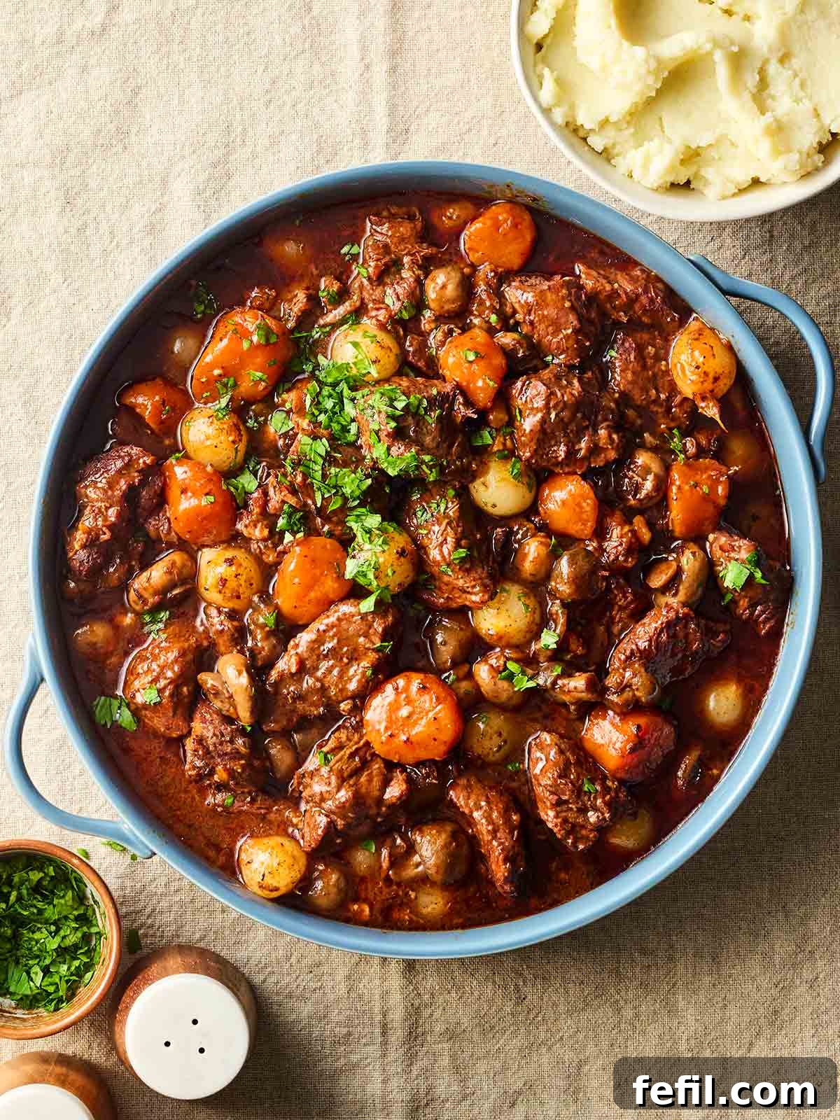 A big pan of beef bourguignon with a bowl of mashed potatoes to the side, on a table, ready to serve, inviting a hearty meal.