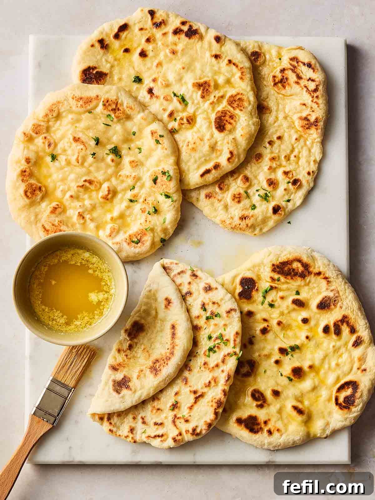 A white wooden board displaying small, round, homemade flatbreads, some brushed with melted butter on the side.