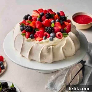 A homemade pavlova dessert, on a cake stand, topped with whipped cream and fresh berries, with raspberry sauce in the background.