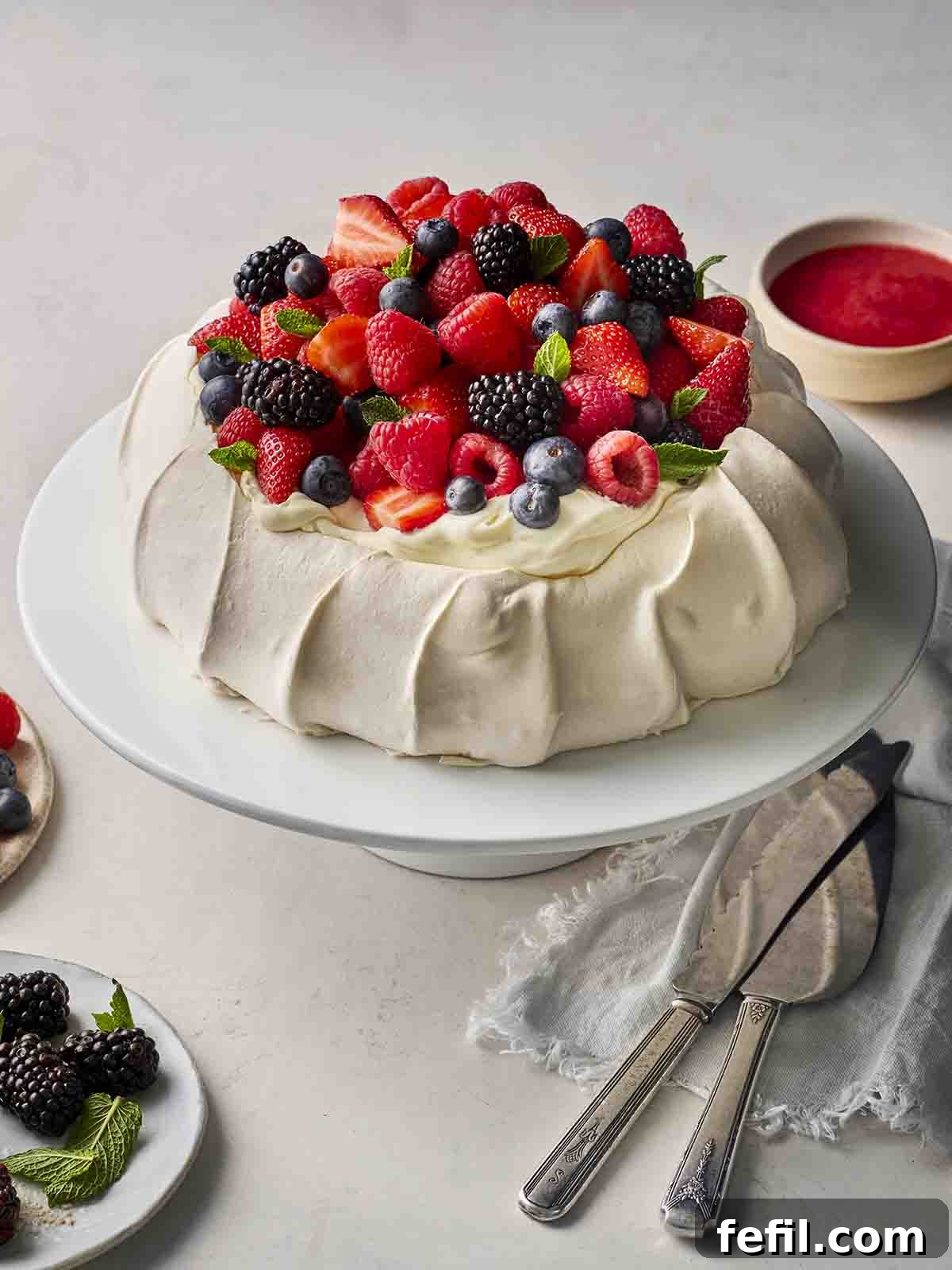 Perfect Pavlova, Made Simple 3 A beautifully assembled Pavlova on a cake stand, adorned with whipped cream, fresh berries, and raspberry sauce, ready to be served at a summer gathering.
