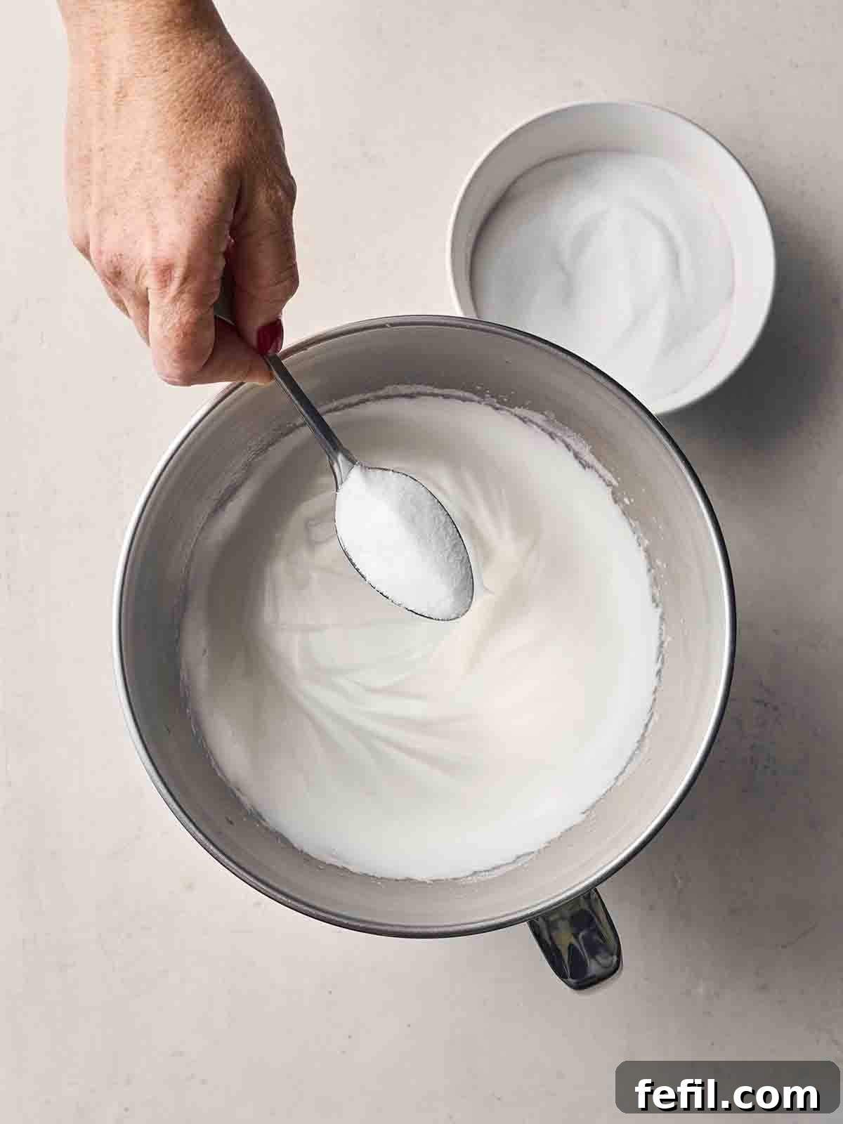 Perfect Pavlova, Made Simple 5 A close-up shot of egg whites being whisked in a large mixing jug, with a hand poised to add caster sugar, illustrating the start of the meringue-making process.