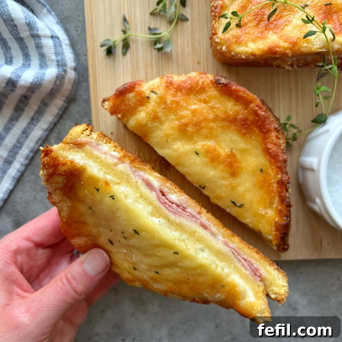 Golden Baked Ham and Cheese Sandwich 2 A hand holds half of a toasted Croque Monsieur filled with savory ham and decadent melted cheese, presented on a rustic wooden board alongside fresh green herbs and an elegant striped cloth napkin, showcasing its gourmet appeal.