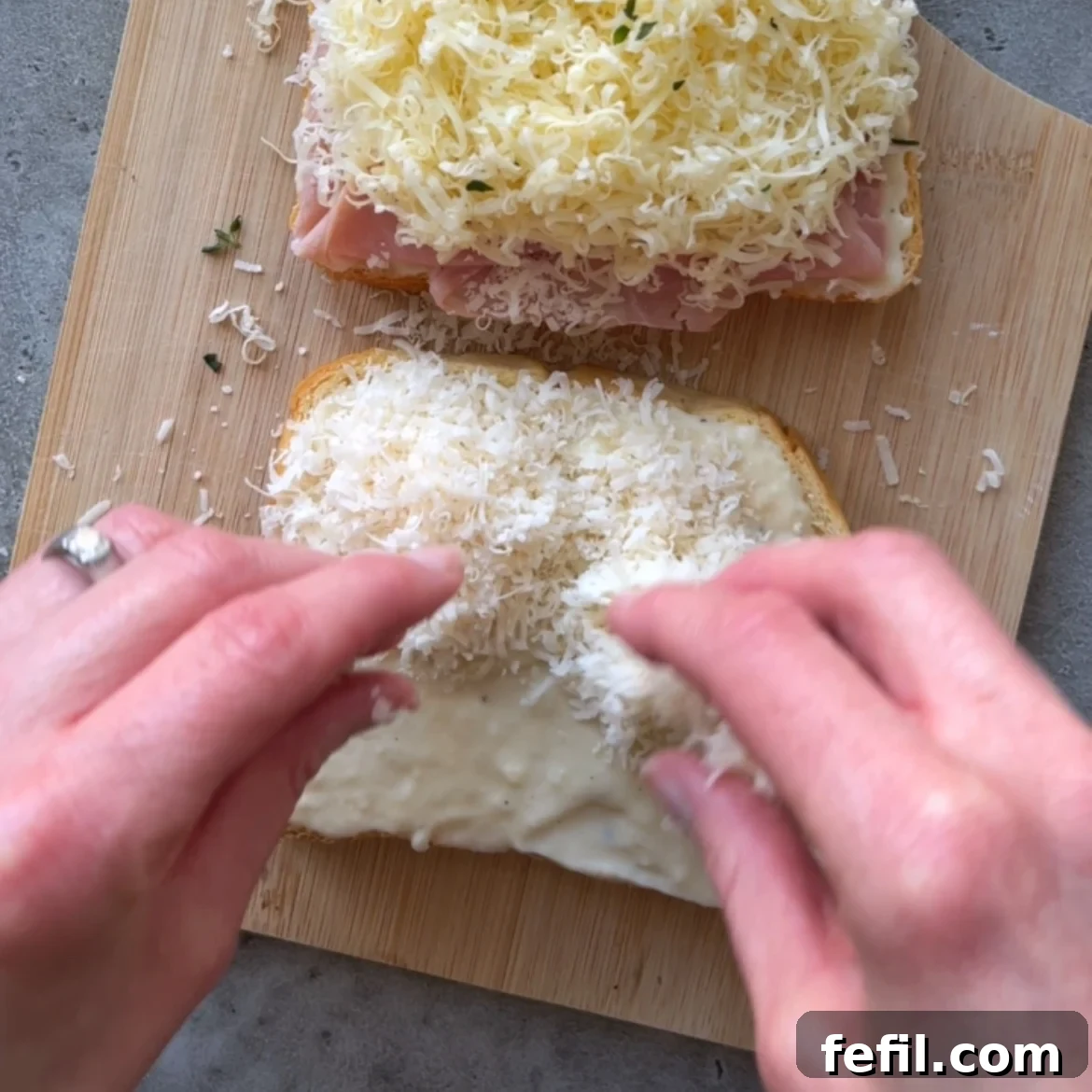 Golden Baked Ham and Cheese Sandwich 7 A pair of hands delicately sprinkling a generous amount of freshly grated cheese onto a slice of bread already coated with creamy béchamel, a key step in assembling a classic Croque Monsieur before baking.