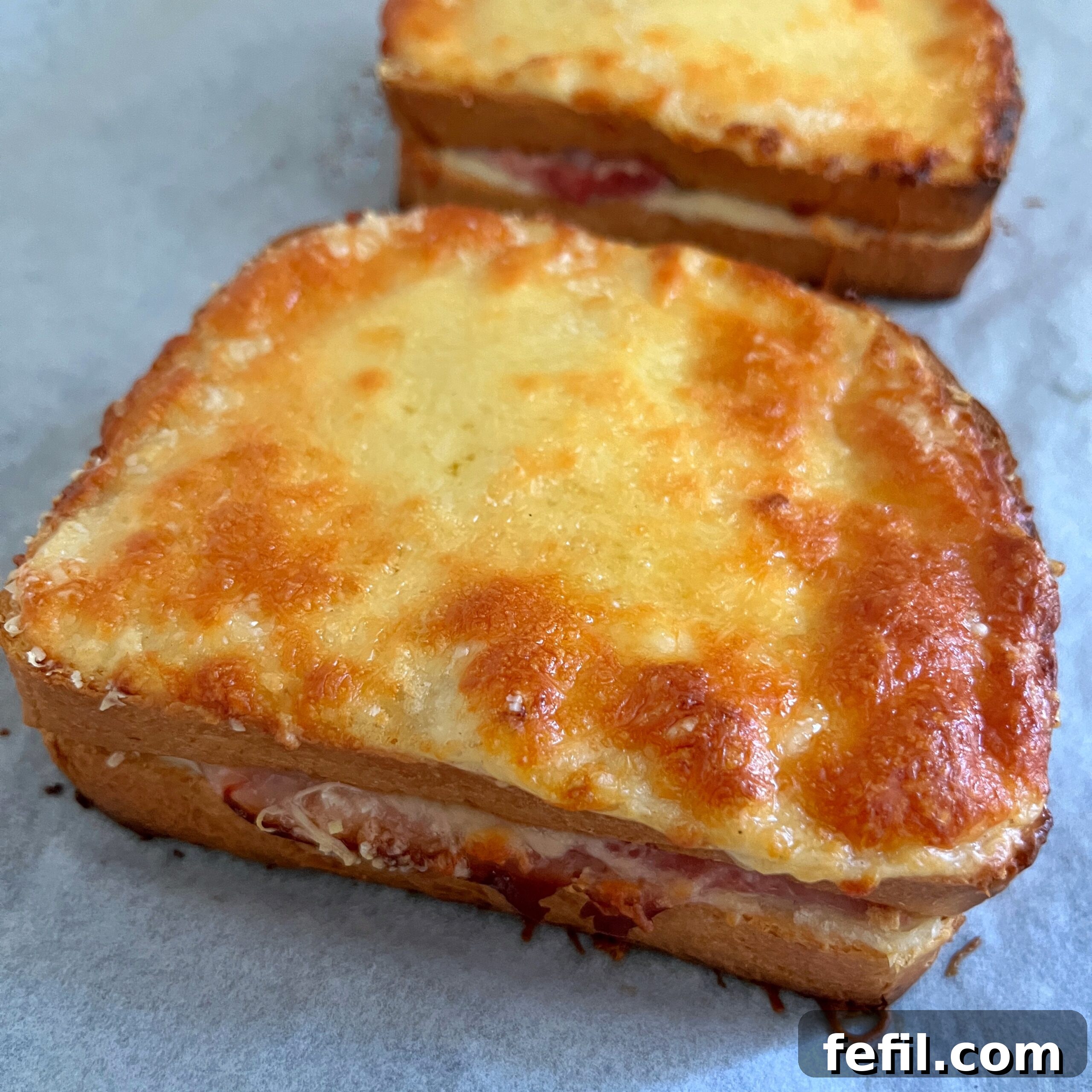 Golden Baked Ham and Cheese Sandwich 8 Two freshly baked Croque Monsieur sandwiches, featuring a perfectly melted, golden-brown cheese top, resting on parchment paper on a baking sheet, ready to be served and enjoyed.
