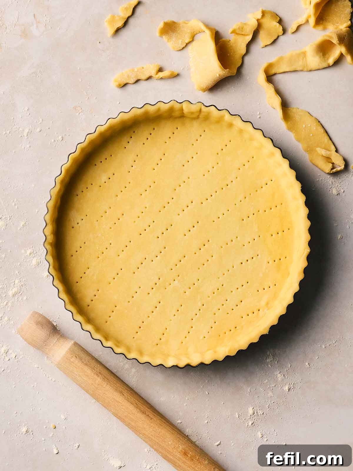 A flan dish with unbaked homemade shortcrust pastry inside with a rolling pin to the side and offcuts of pastry, on a white surface.